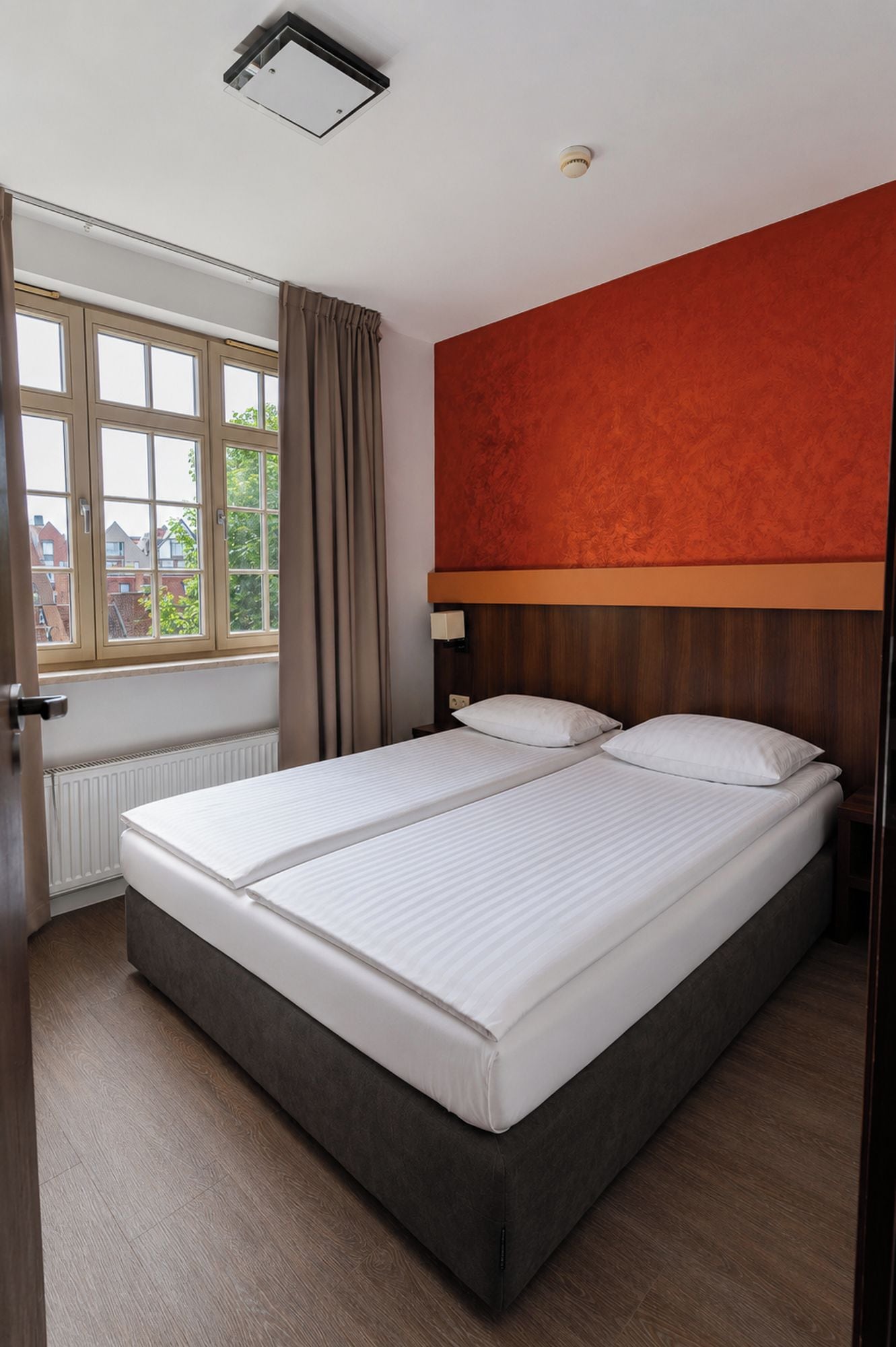 A hotel room with a large bed, a window with curtains, a red accent wall, and a ceiling light.