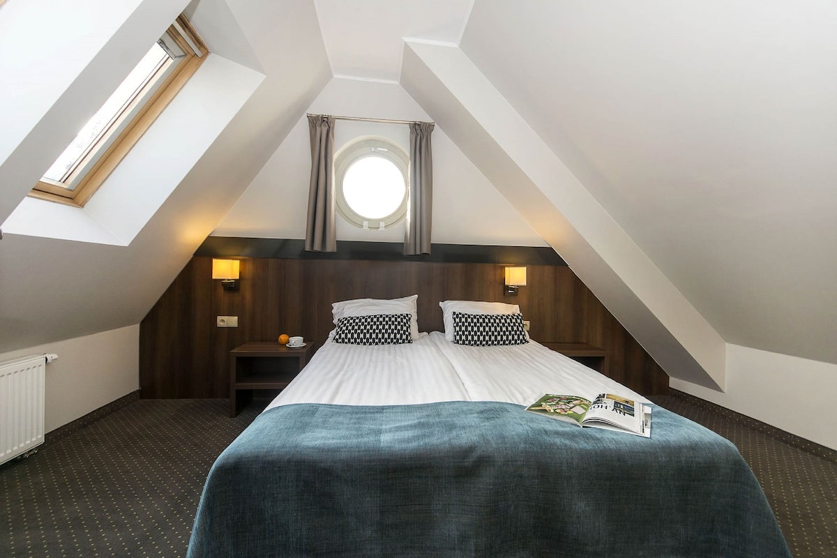 Une chambre avec une lucarne, une fenêtre ronde, deux tables de chevet avec des lampes, un lit recouvert d’une couverture bleue et un magazine posé sur le lit.