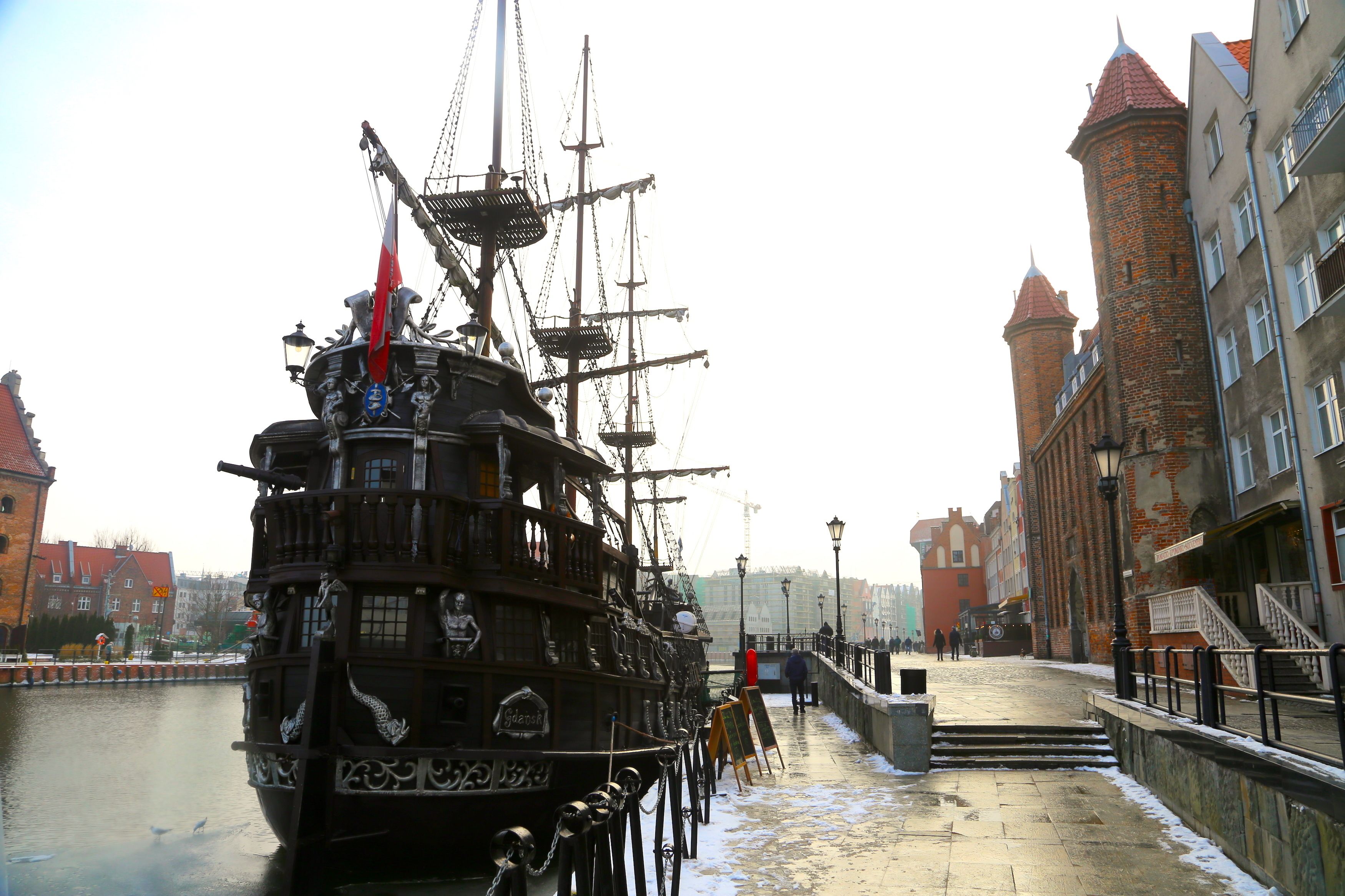 A historic ship docked at a canal with buildings and a bridge in the background.