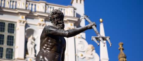 Una estatua de figura musculosa sosteniendo una lanza, frente a un edificio clásico con detalles ornamentales y un cielo azul despejado.