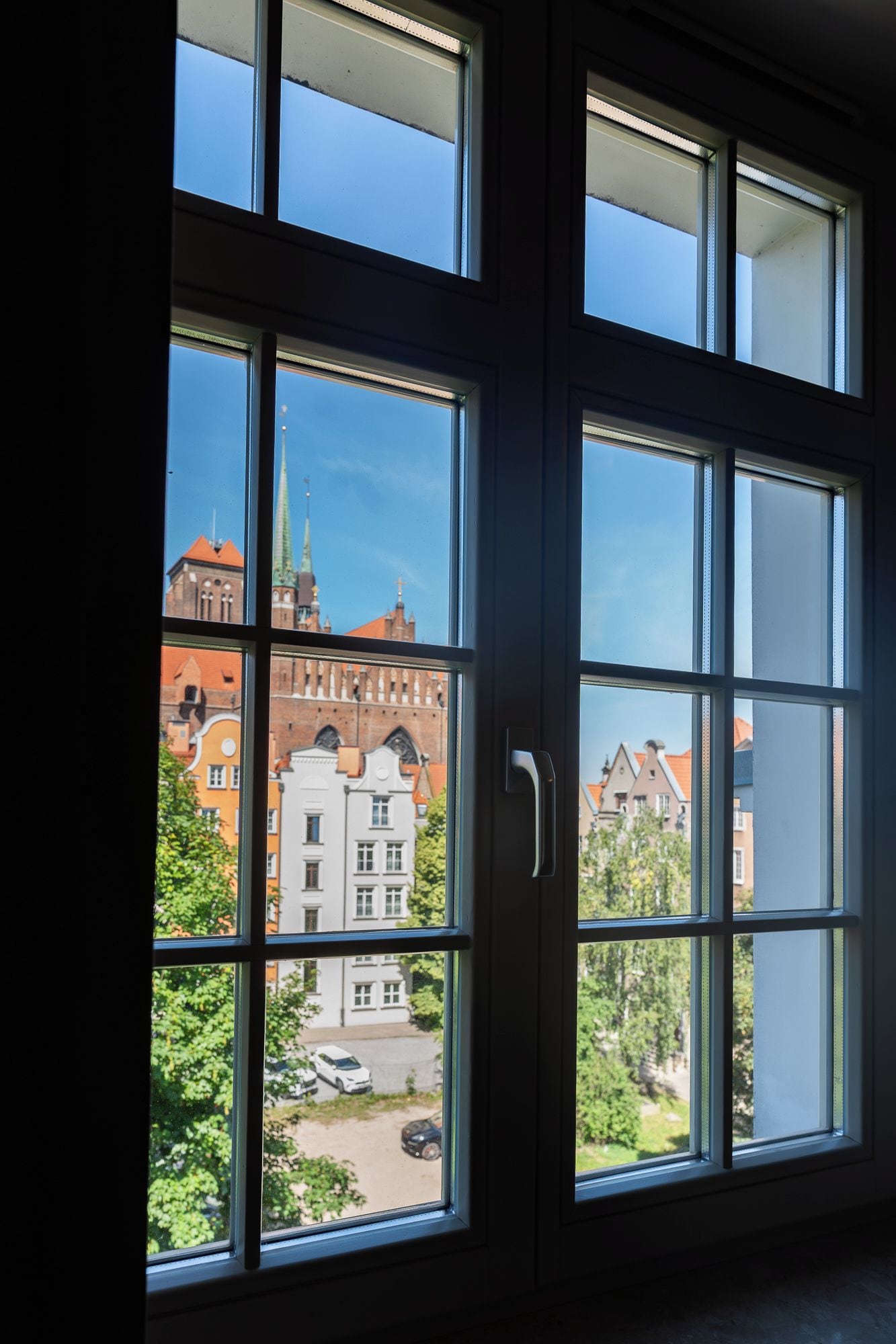 Une vue à travers une fenêtre à carreaux multiples, révélant un bâtiment avec un toit rouge et une flèche, entouré de verdure et de voitures garées.