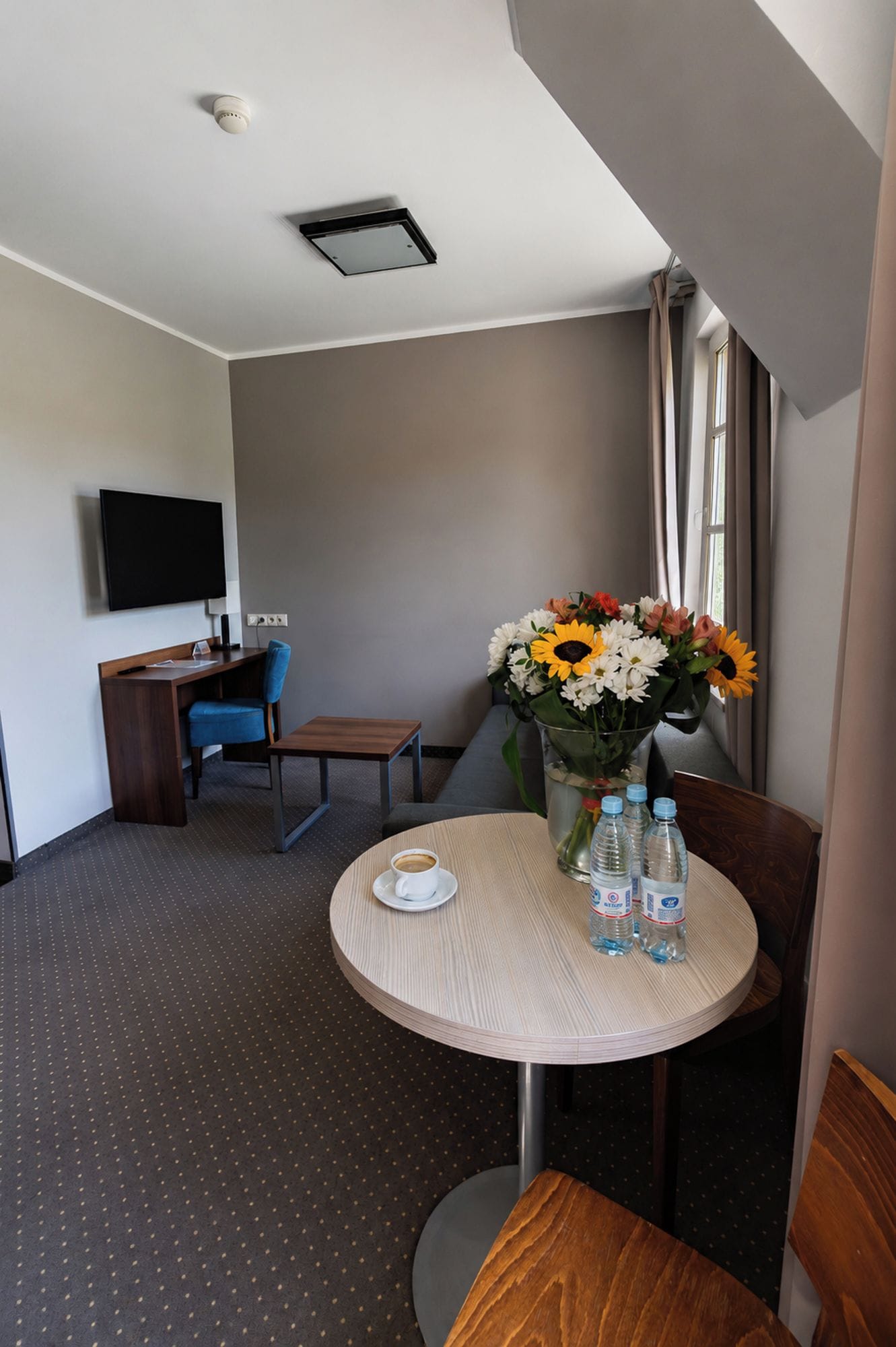 A hotel room with a round table, a chair, a desk with a chair, a flat-screen TV, and a window with curtains.