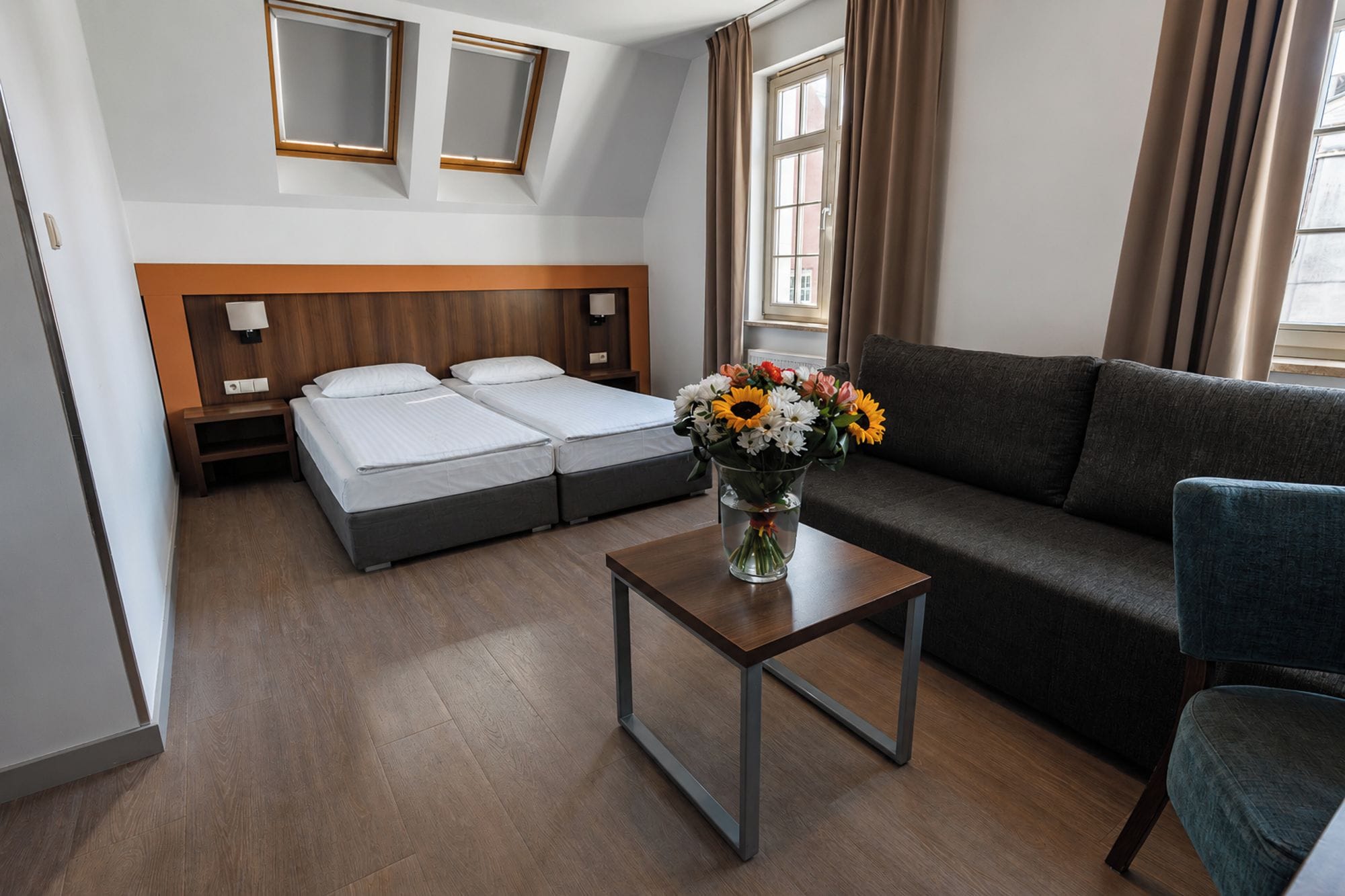 A hotel room with a double bed, a sofa, a small table with a vase of flowers, and a window with curtains.