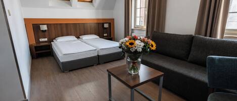 A hotel room with a double bed, a sofa, a small table with a vase of flowers, and a window with curtains.