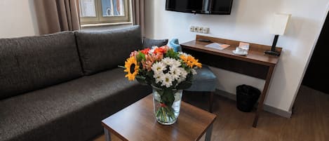 Quarto de hotel com sofá, mesinha com um vaso de flores, escrivaninha com controle remoto e televisão de tela plana fixada na parede.