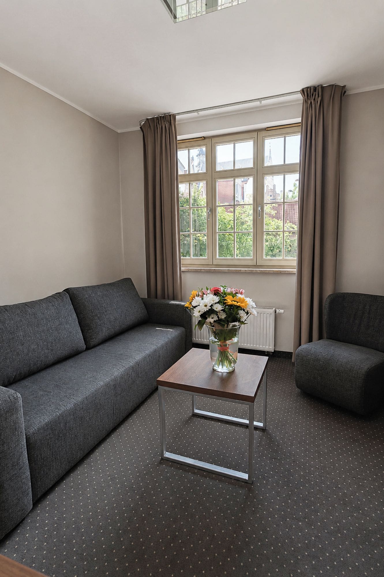 A modern living room with a grey sectional sofa, a clear glass coffee table with a vase of flowers, and a window with curtains.