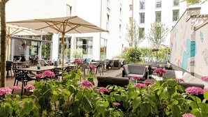 An outdoor patio area with tables and chairs, a large umbrella, and a sofa. - Motel One Köln-Mediapark (Cologne)