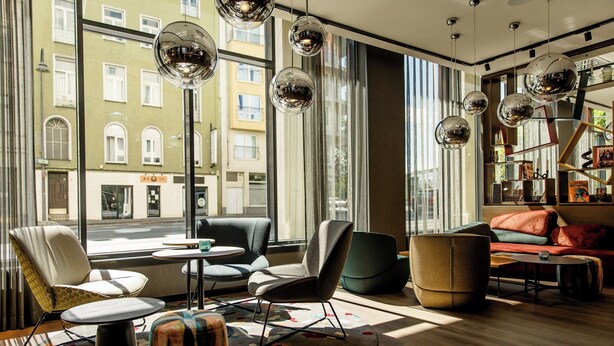 A modern interior with large windows, a sofa, chairs, and a coffee table. - Motel One Köln-Mediapark (Cologne)