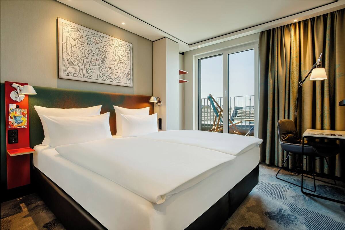 A hotel room with a large bed, a desk with a chair, a window with curtains, and a painting on the wall.