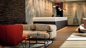 A modern lobby with a reception desk, a red armchair, a beige sofa, and a round coffee table. - Motel One Köln-Mediapark (Cologne)