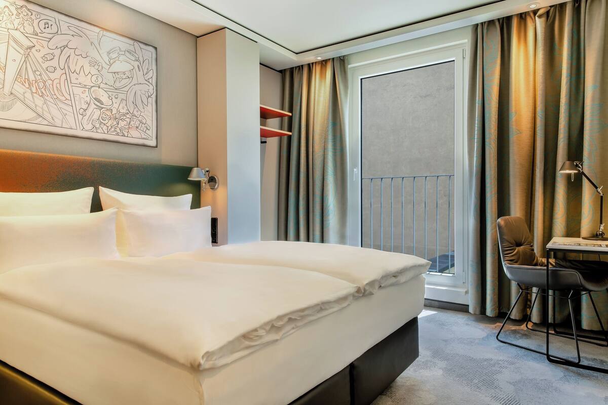 A hotel room with a large bed, a desk with a chair, a window with curtains, and a wall-mounted artwork.
