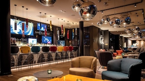 A modern bar area with a variety of seating options, including round tables and chairs, and a bar counter with a selection of bottles. - Motel One Köln-Mediapark (Cologne)