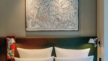 Ein Hotelzimmer mit einem gerahmten, Comic-artigen Kunstwerk an der Wand, einem roten Nachttisch mit Lampe und einem grünen Kopfteil mit zwei weißen Kissen.