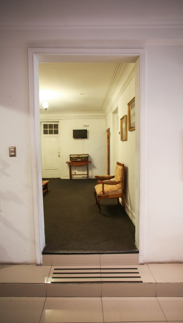A hallway with a carpeted floor, a wooden bench, a chair, a television mounted on the wall, and a door at the end.