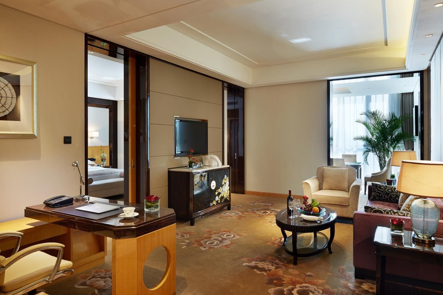 A modern hotel room with a large bed, a desk with a laptop, a television, and a seating area with a sofa and armchairs.