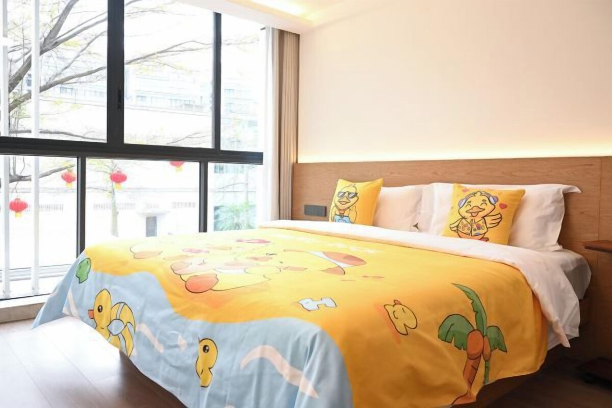 Une chambre d’hôtel avec une grande fenêtre, un lit recouvert d’une courtepointe aux couleurs vives et une tête de lit en bois.