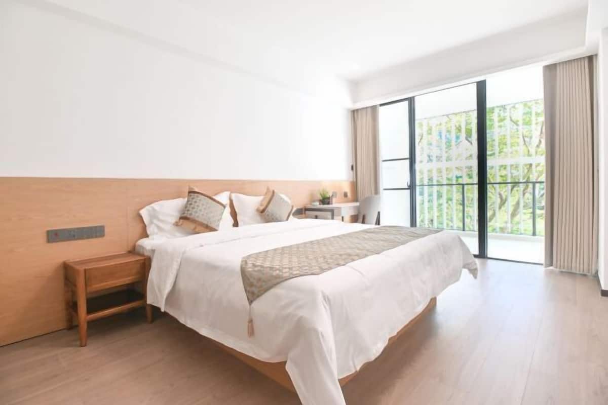 Una habitación de hotel moderna con una cama grande, una mesita de noche de madera, una pequeña mesa y vistas a zonas verdes a través de amplias ventanas.