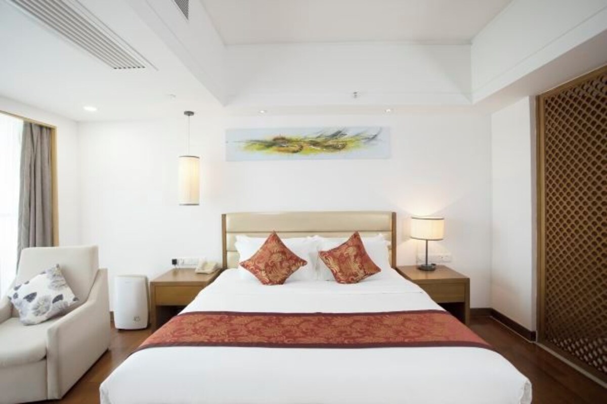 Une chambre d’hôtel avec un lit, deux lampes de chevet, une chaise, un tableau au mur et une fenêtre avec des rideaux.