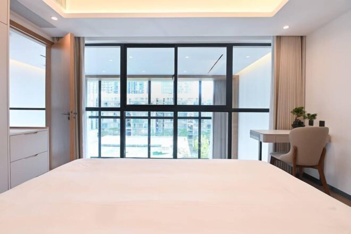 Habitación de hotel con una cama grande, un escritorio, una silla y vistas a edificios a través de amplias ventanas.