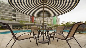 Ein Poolbereich mit Tisch und Stühlen unter einem gestreiften Sonnenschirm, umgeben von einem modernen Gebäude mit Balkonen.