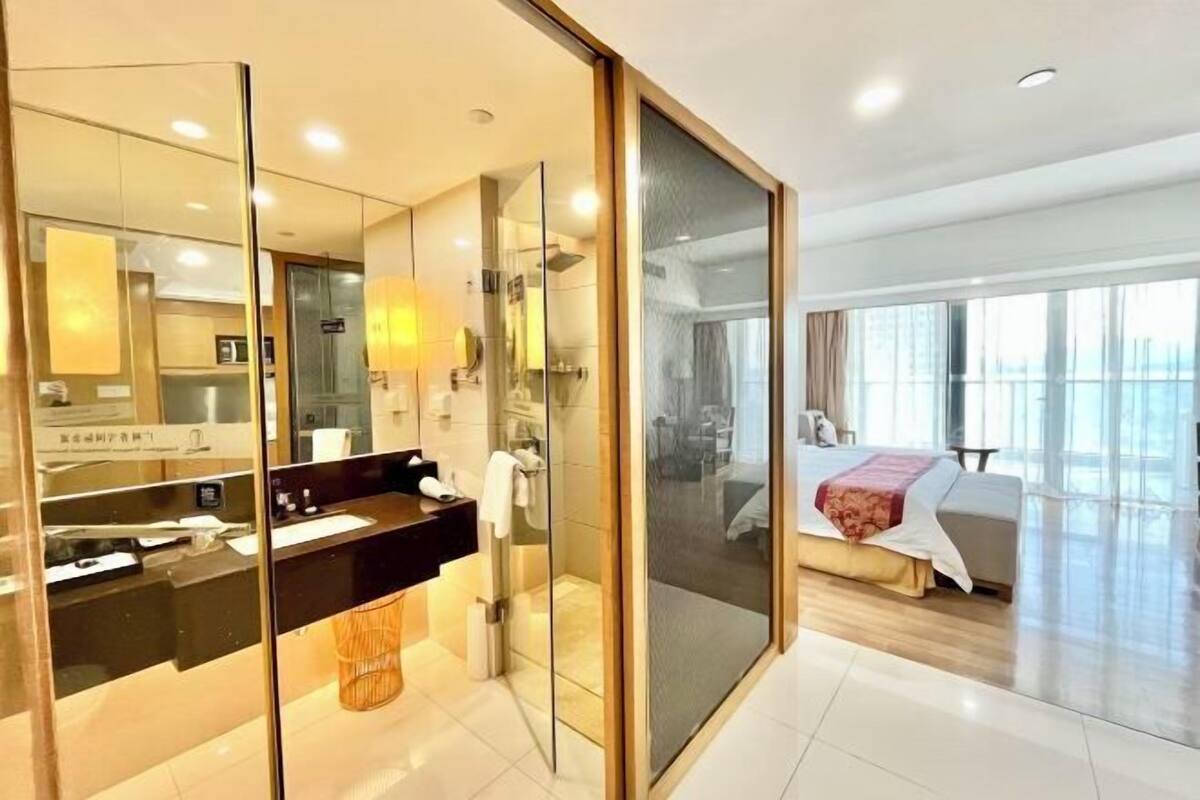 Habitación de hotel moderna con separador de vidrio, cama, baño con ducha de vidrio y vistas a la ciudad.