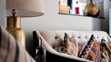 A tufted sofa with patterned cushions, a brass lamp, and a shelf with books and bottles.