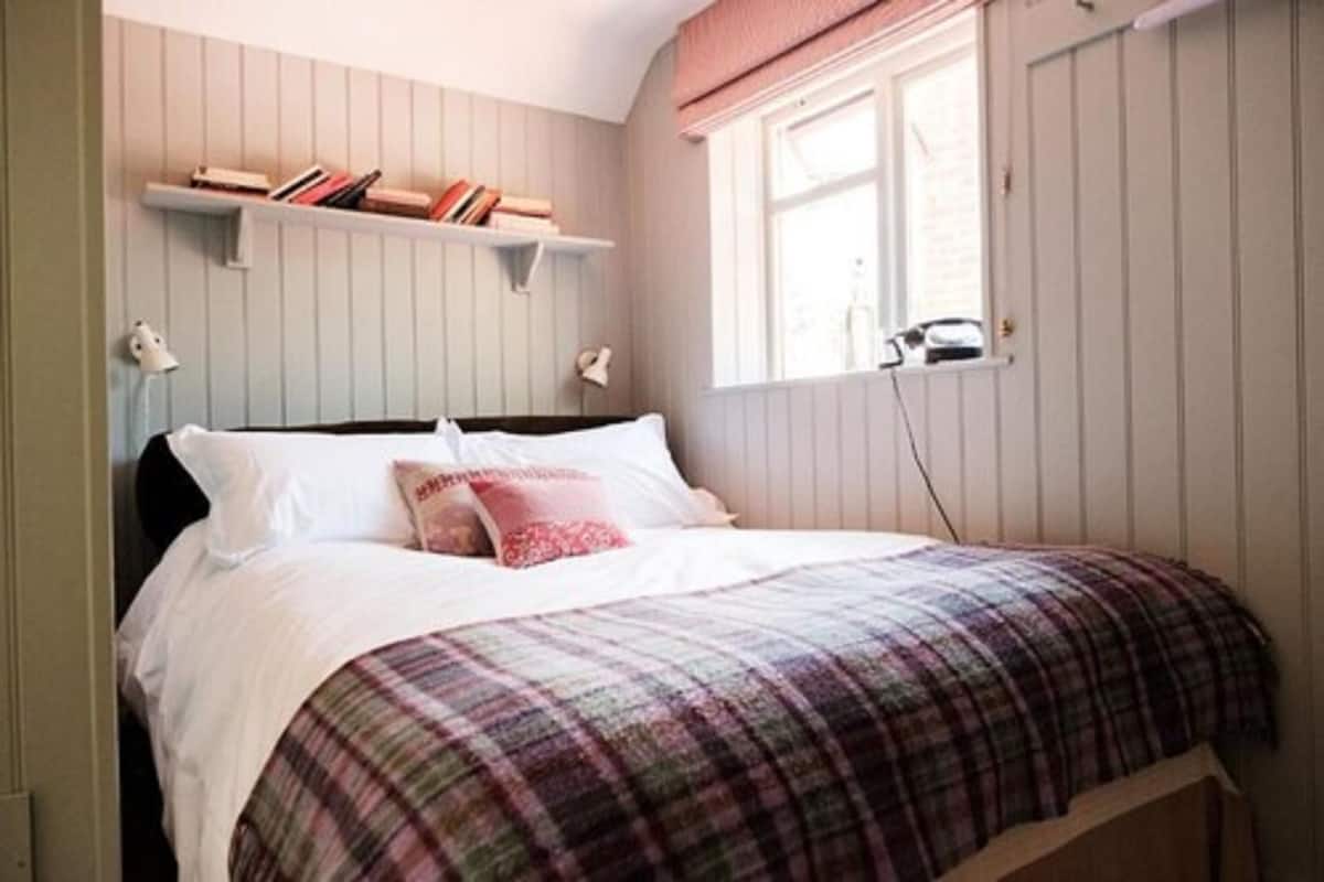 A bedroom with a bed, a window with blinds, a shelf with books, and a bedside lamp.