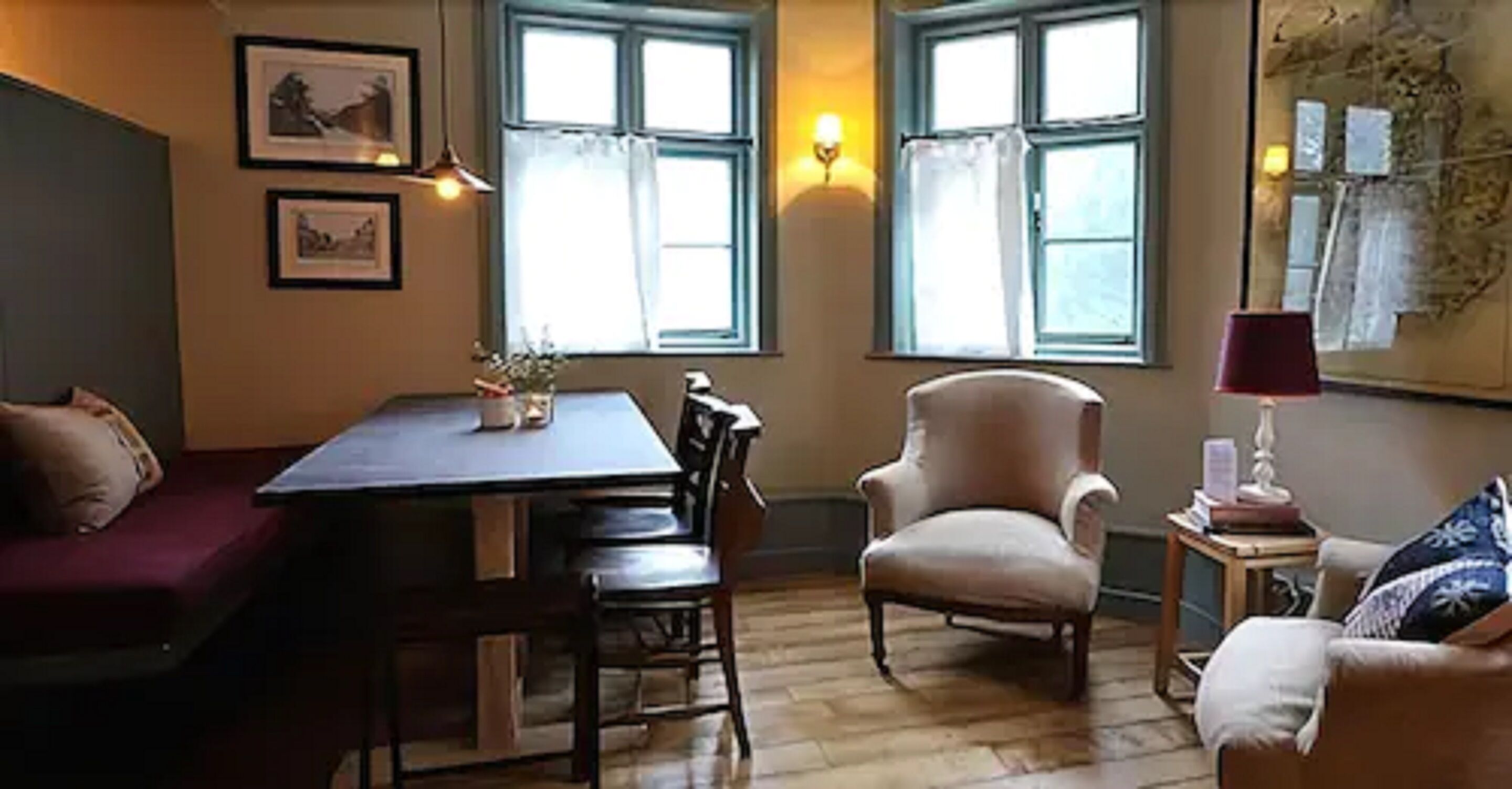 A cozy room with a wooden floor, a dining table with chairs, a cushioned armchair, and a sofa. There are two windows with curtains, a wall-mounted lamp, and framed pictures.
