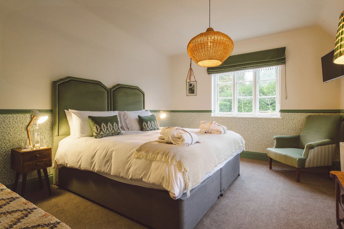 A neatly arranged bedroom with a large bed, a bedside table with a lamp, a chair, and a window with a view of greenery.