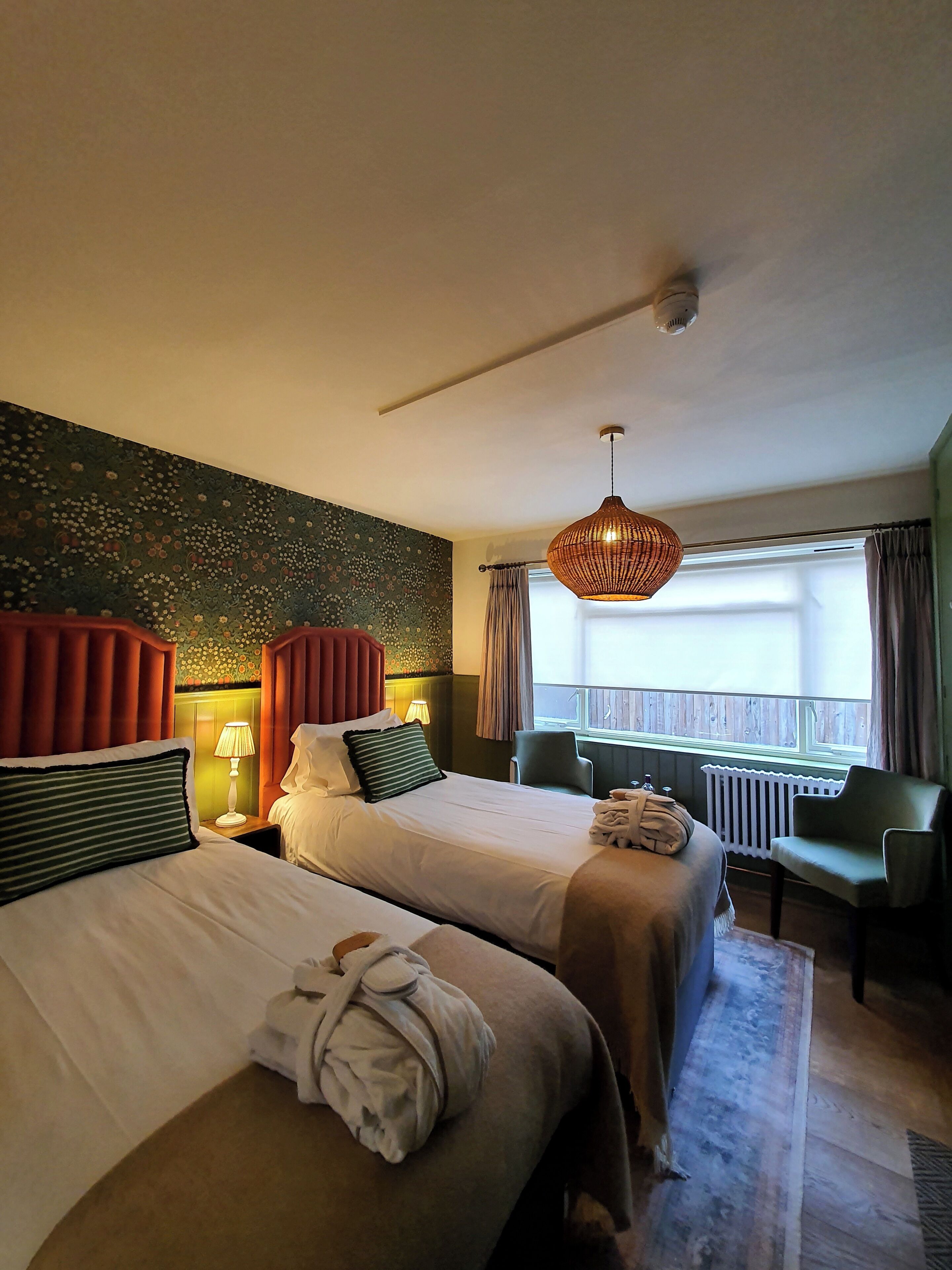 A hotel room with two beds, a sitting area with a chair, a window with curtains, and a hanging light fixture.
