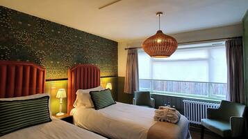 A hotel room with two beds, a sitting area with a chair, a window with curtains, and a hanging light fixture.