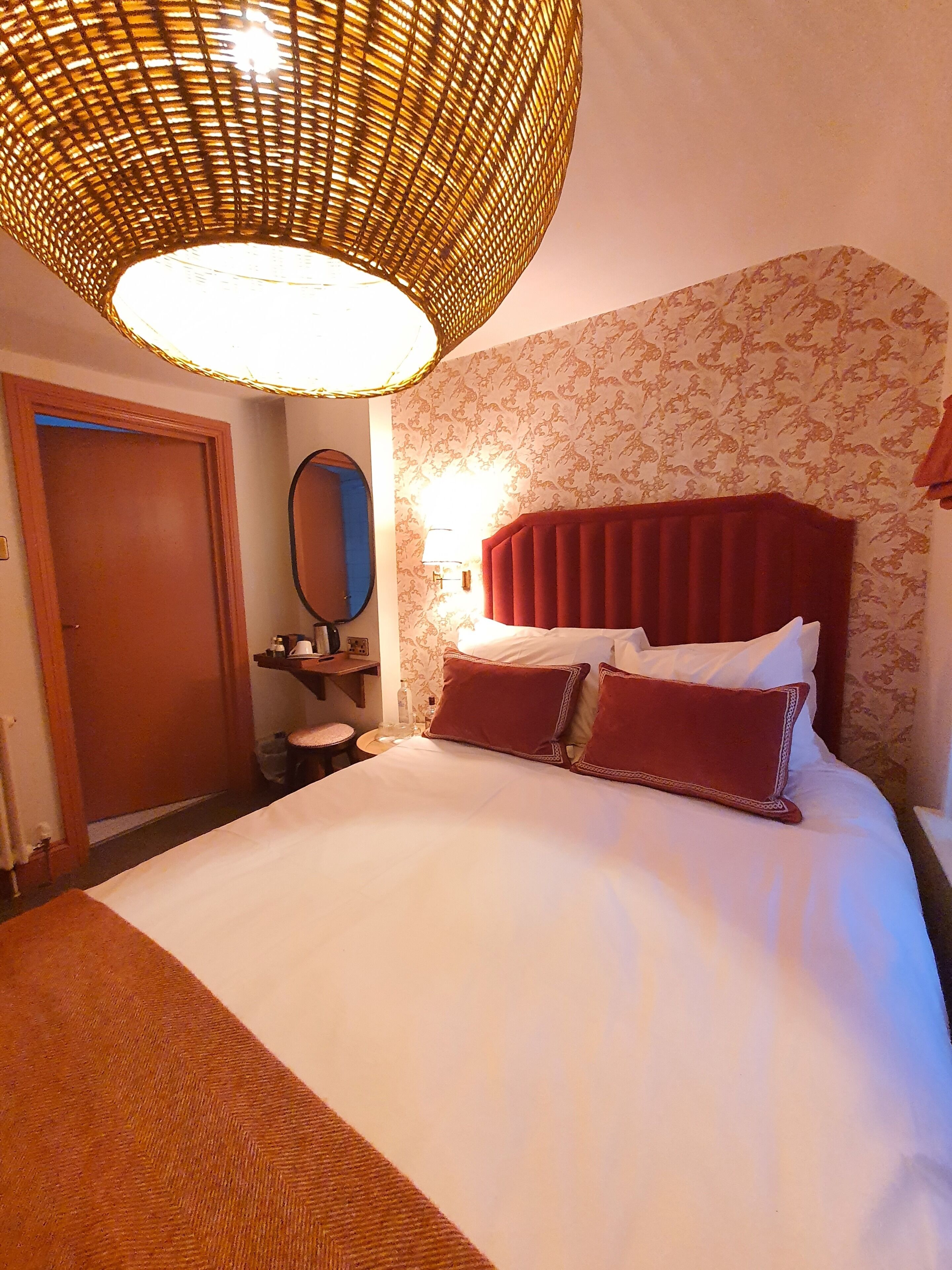 A hotel room with a large bed, a red upholstered headboard, a wall-mounted mirror, and a wicker ceiling light fixture.