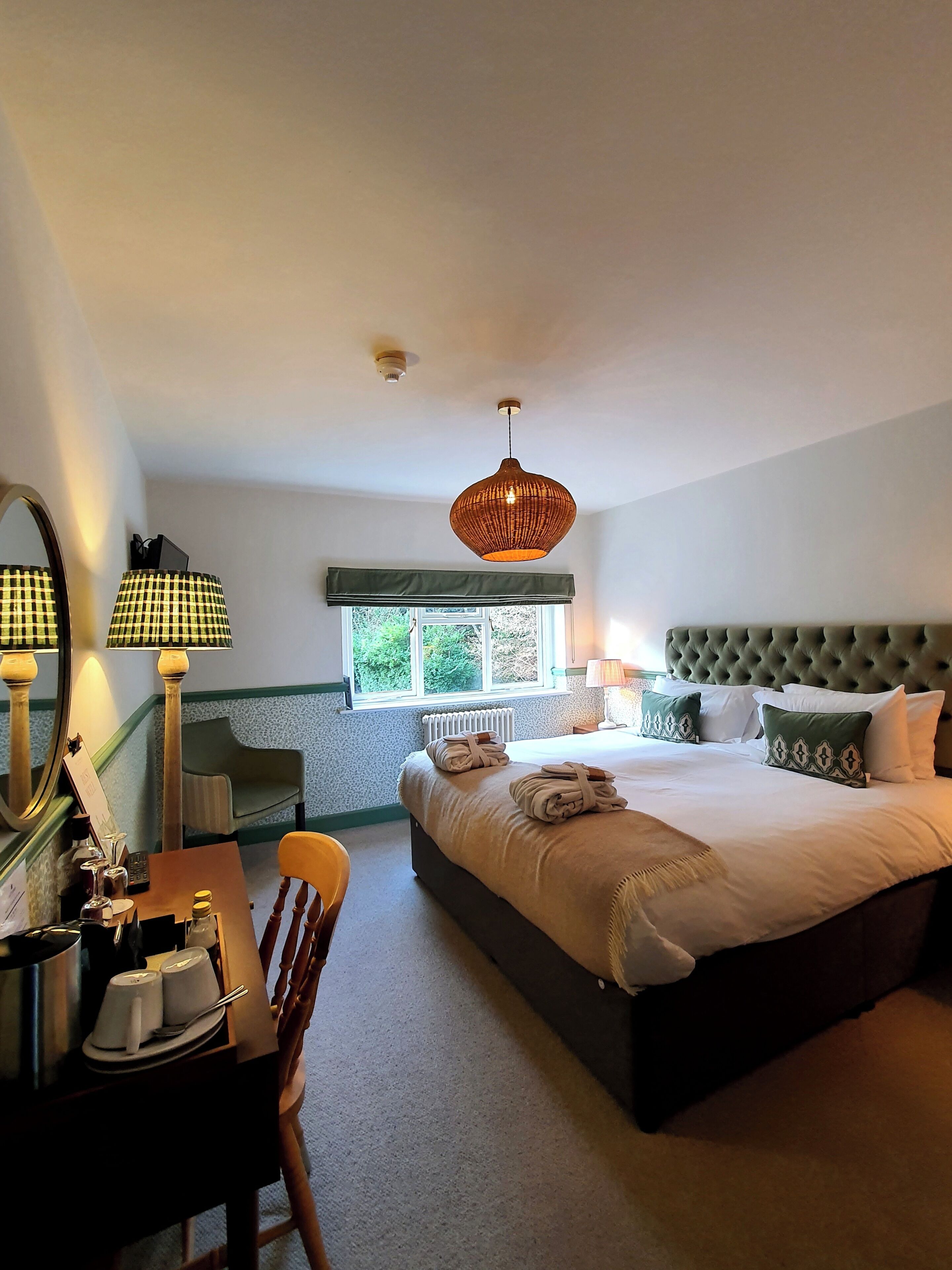A hotel room with a large bed, a bedside table with a mirror, a chair, and a window with a view of greenery.