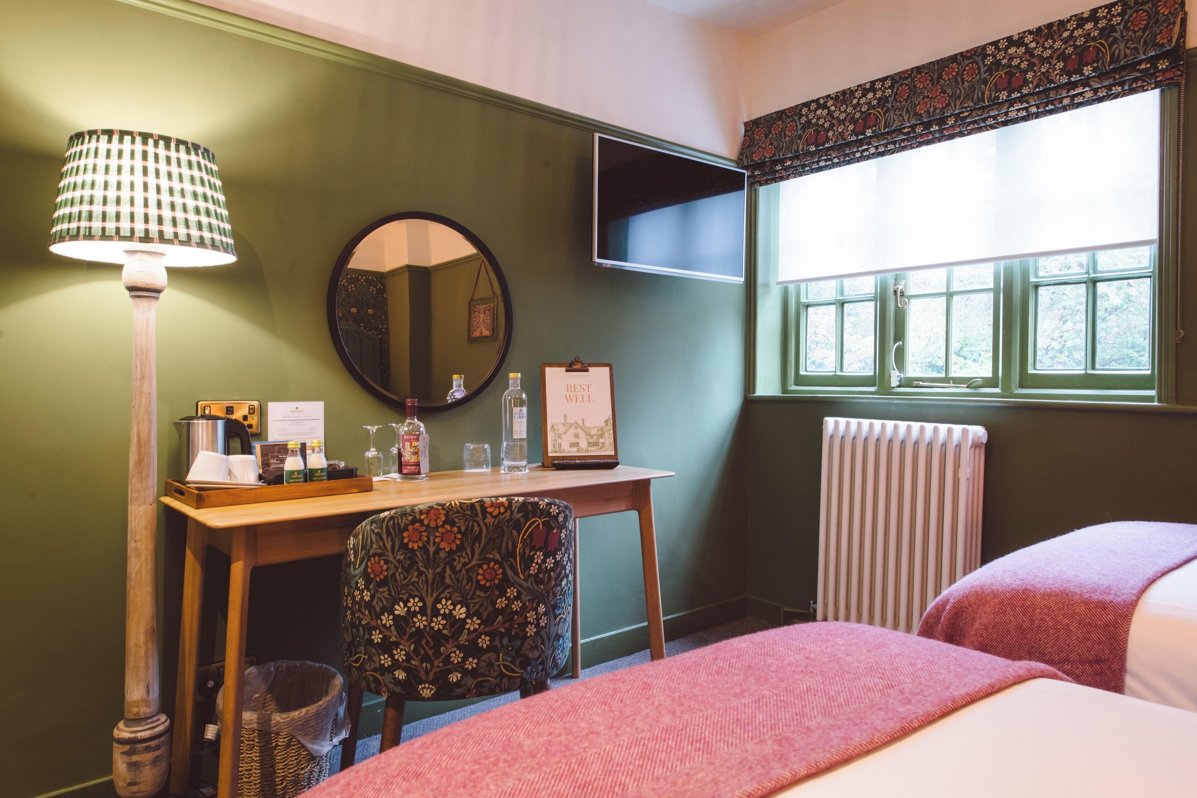 A room with a wooden desk, a mirror, a framed certificate, a radiator, and a window with a floral valance.
