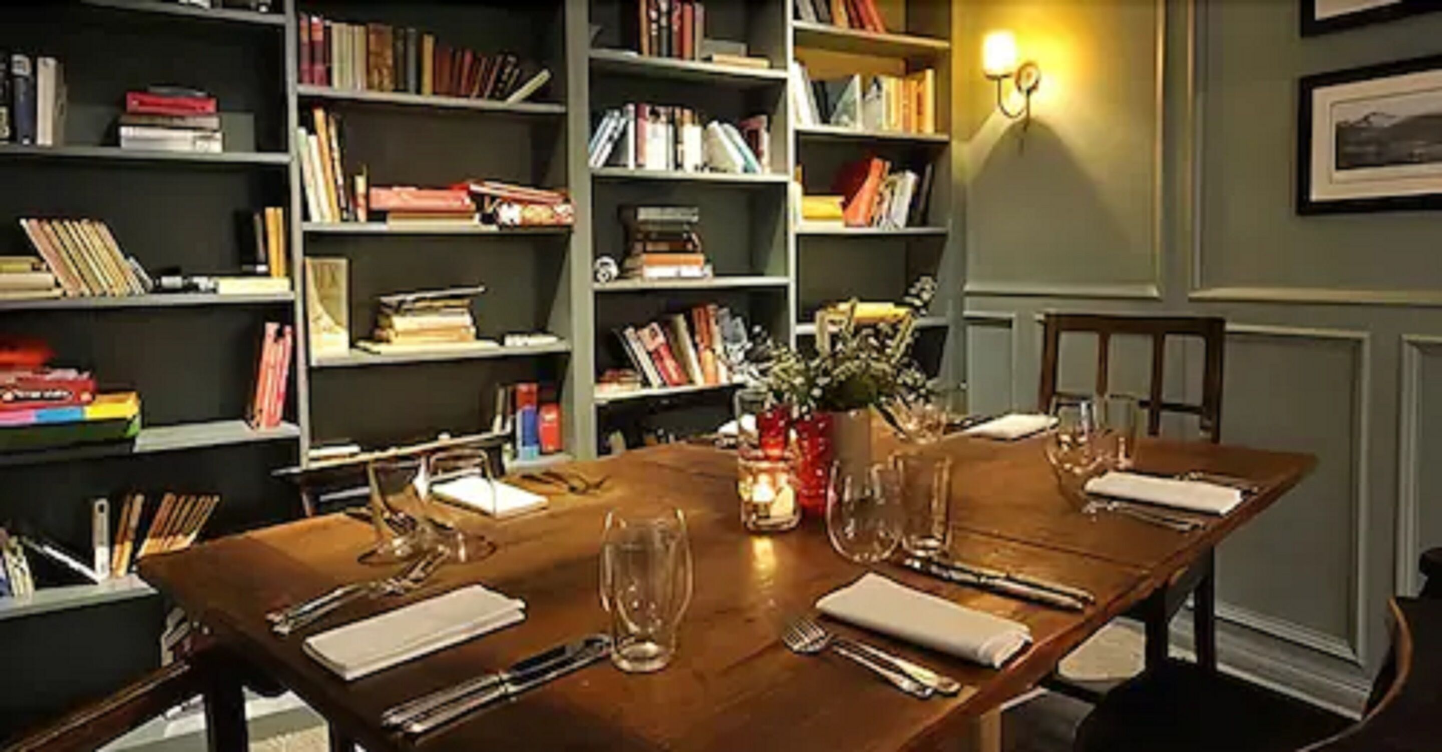 Mesa de comedor preparada para cuatro personas, con un jarrón de flores y rodeada de estanterías llenas de libros.