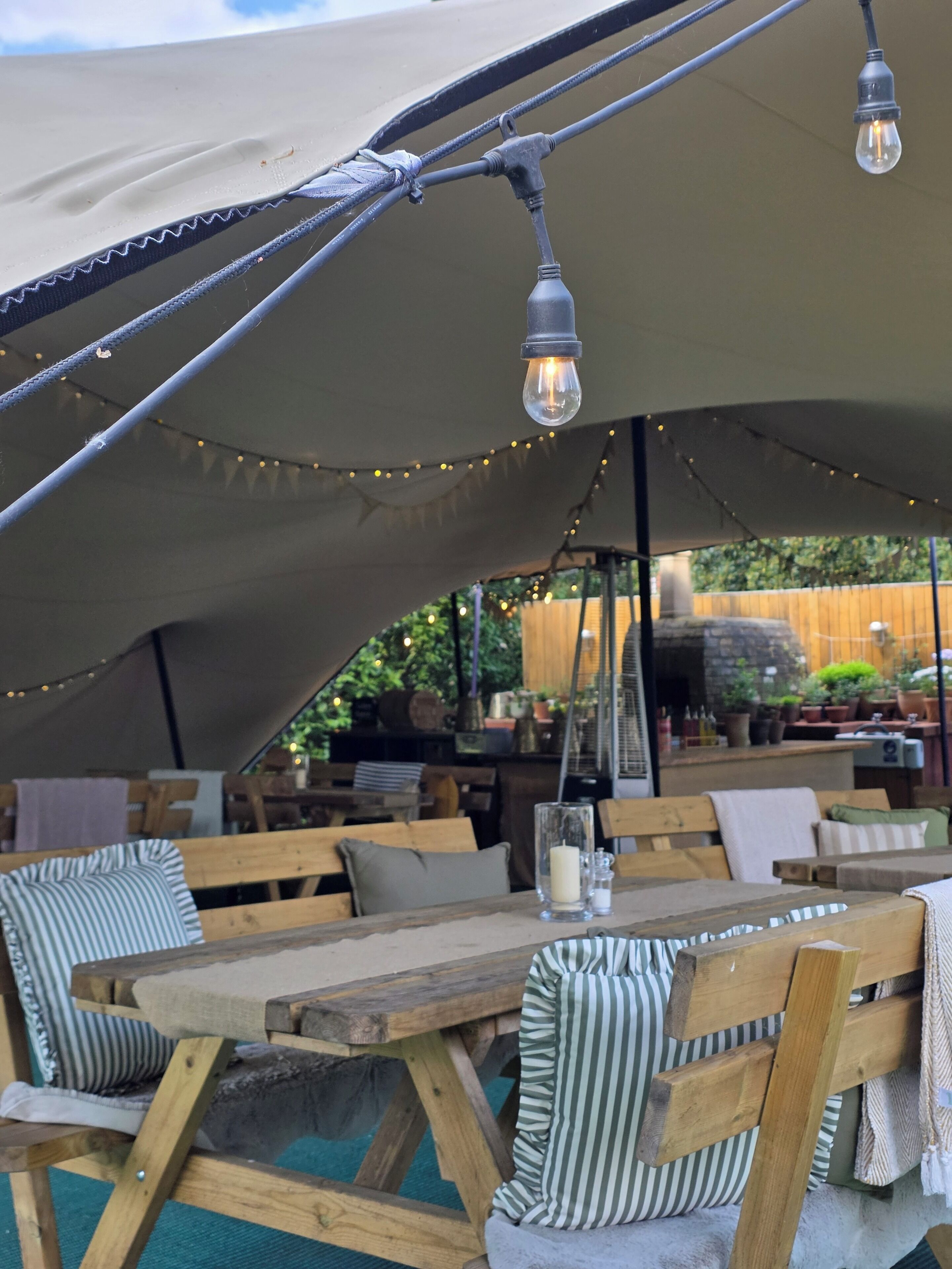 An outdoor dining area with wooden tables and benches, string lights, and a canopy overhead.