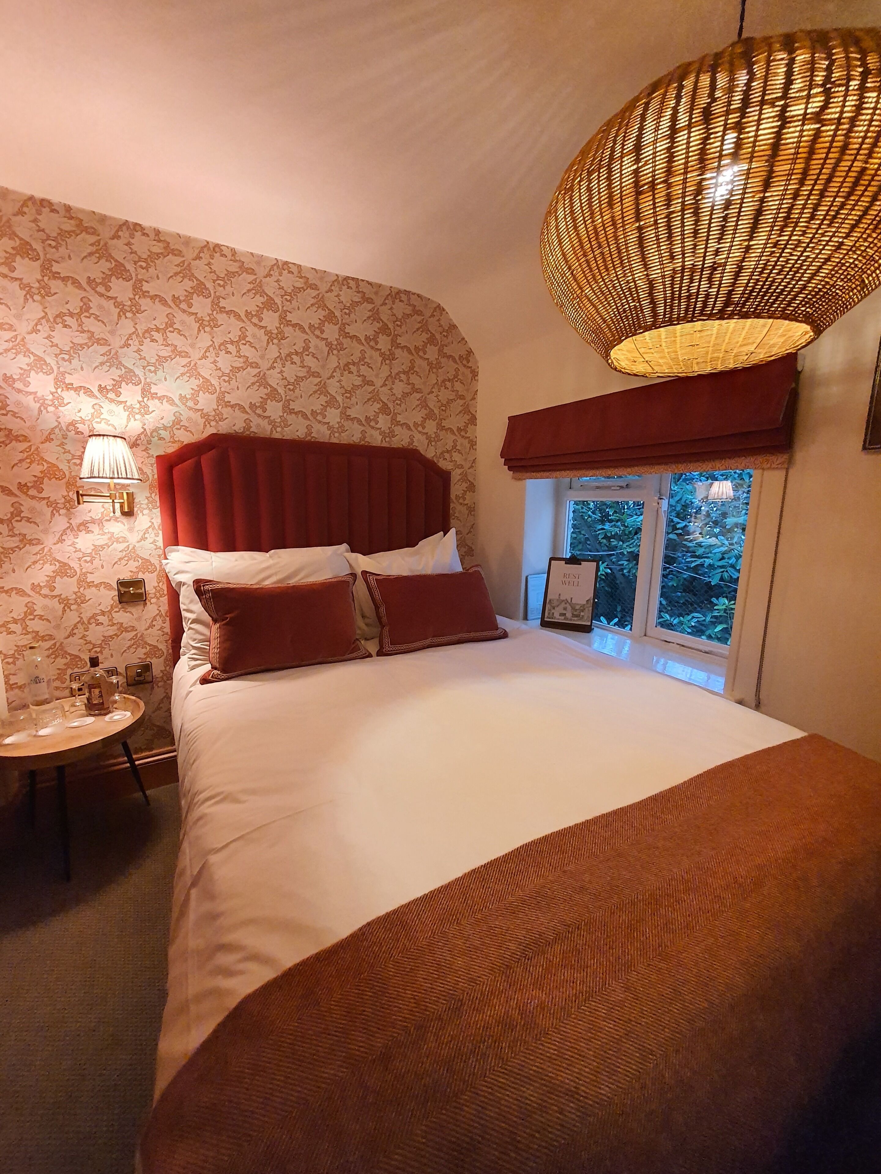 Une chambre d’hôtel avec un grand lit, une table ronde sur laquelle se trouvent une bouteille et des verres, une lampe fixée au mur et une fenêtre donnant sur un paysage verdoyant.
