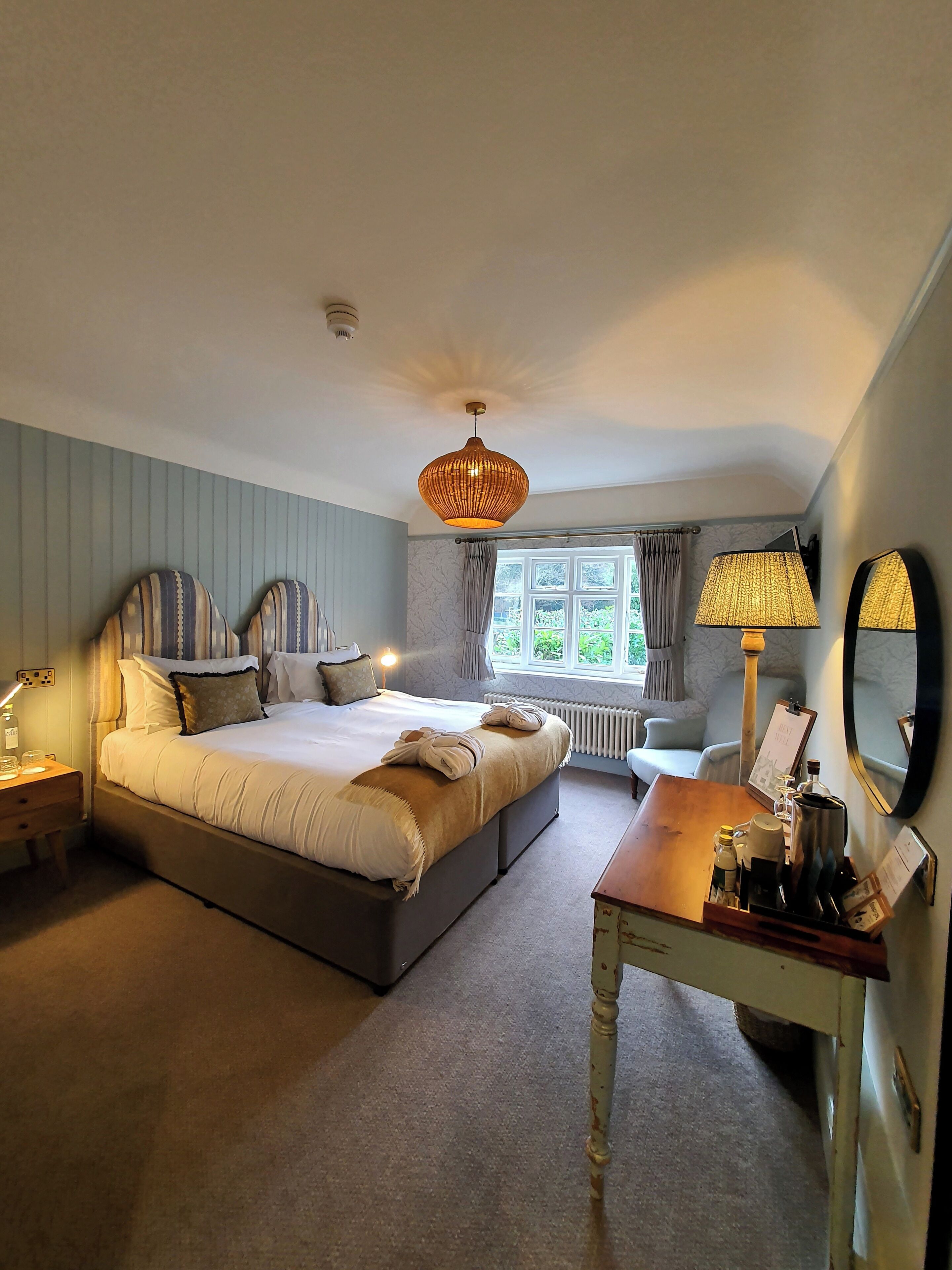 A bedroom with a bed, bedside tables, a desk with a lamp, a window, and a radiator.