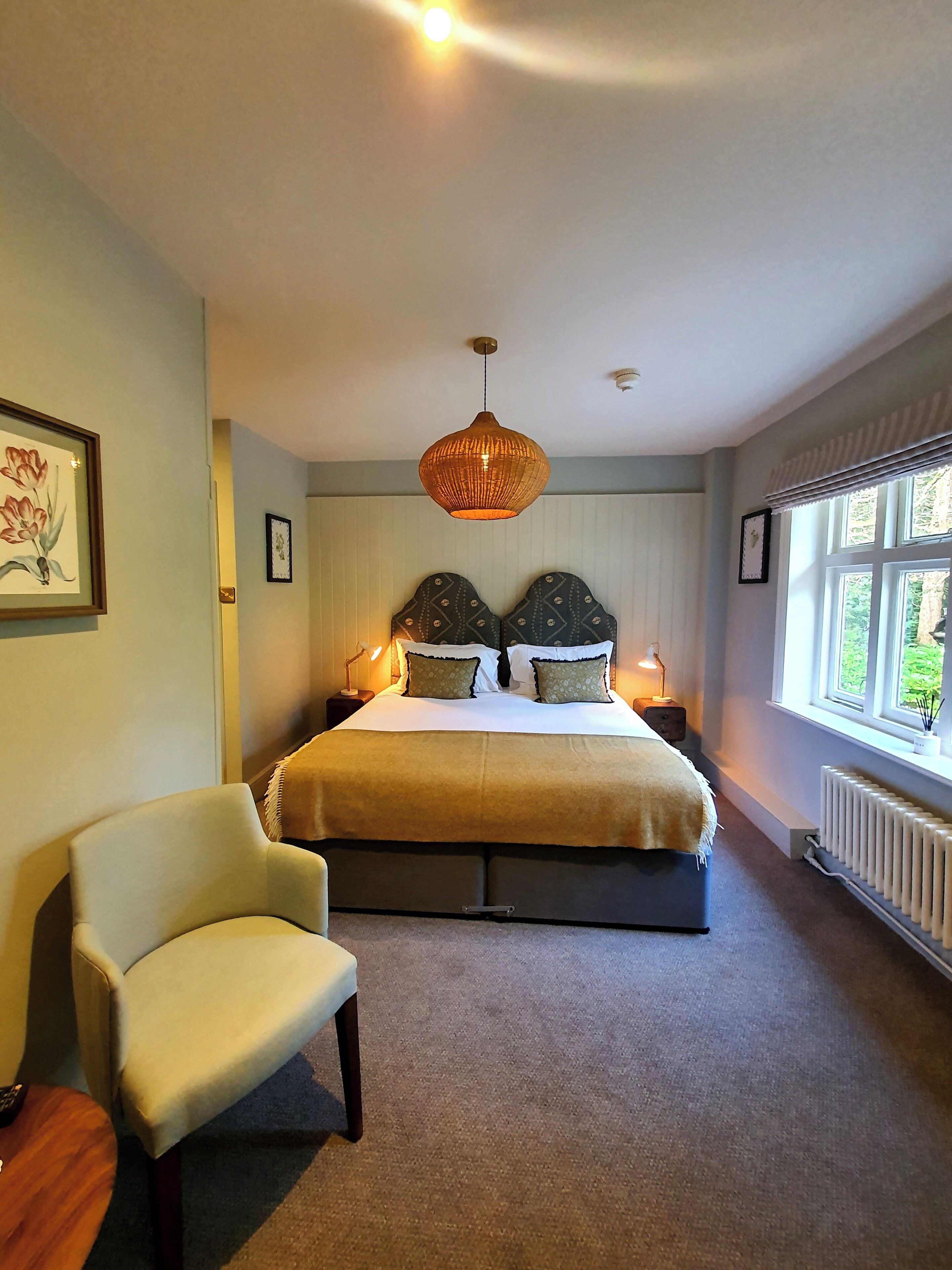 A bedroom with a bed, two bedside lamps, a chair, a hanging light fixture, and a window with a view.