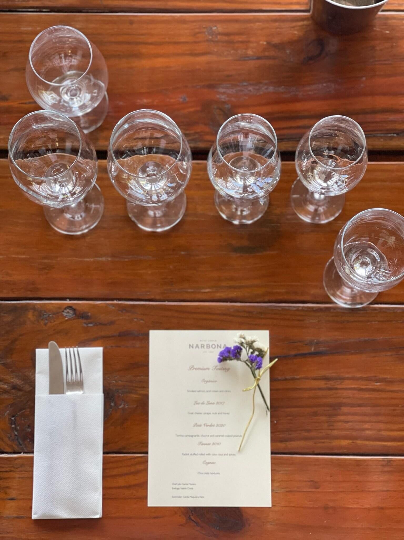 Una mesa de madera con seis vasos vacíos, una servilleta doblada con un tenedor y un menú decorado con una flor.