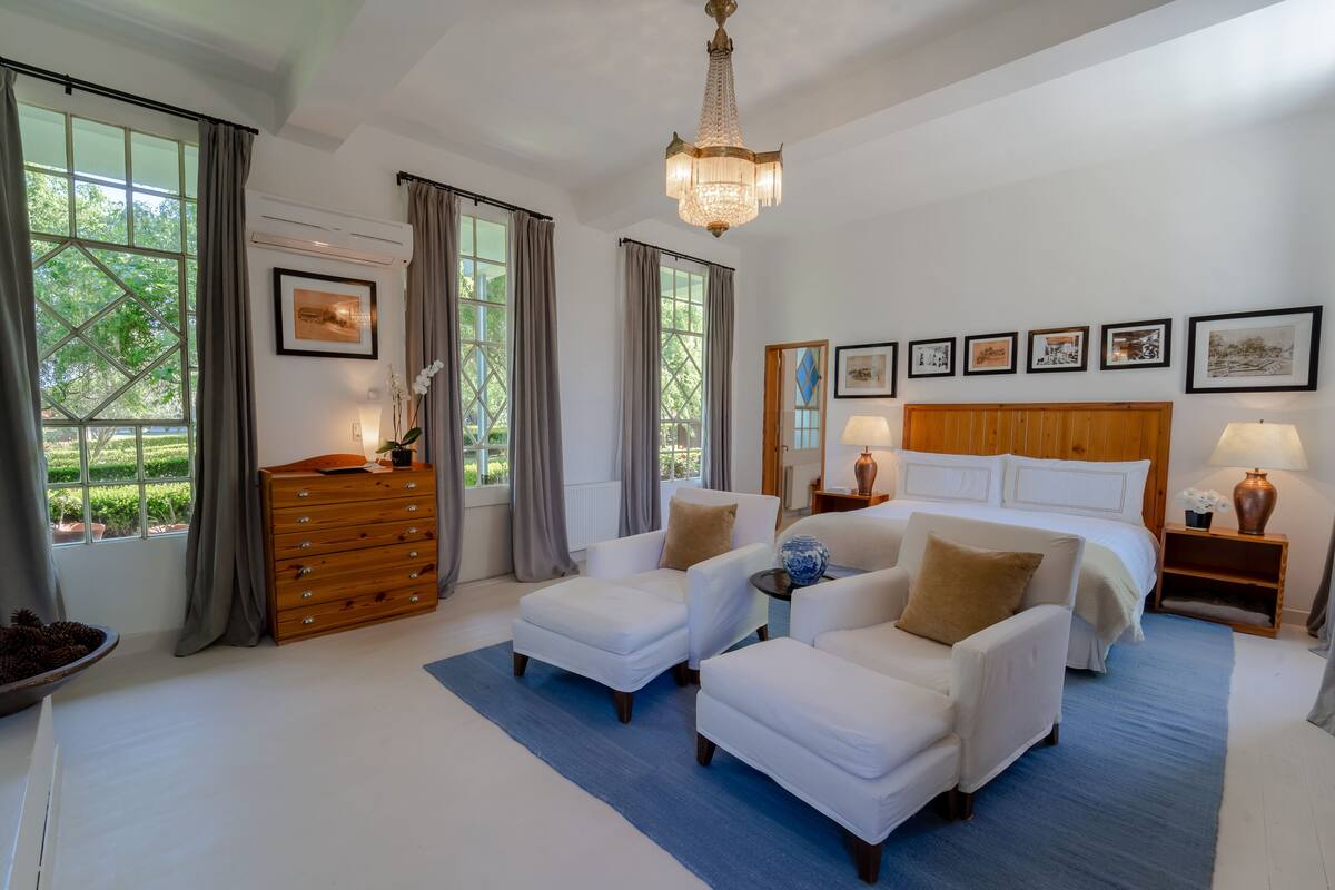 A spacious bedroom with a large bed, two white armchairs, a wooden dresser, and a chandelier.
