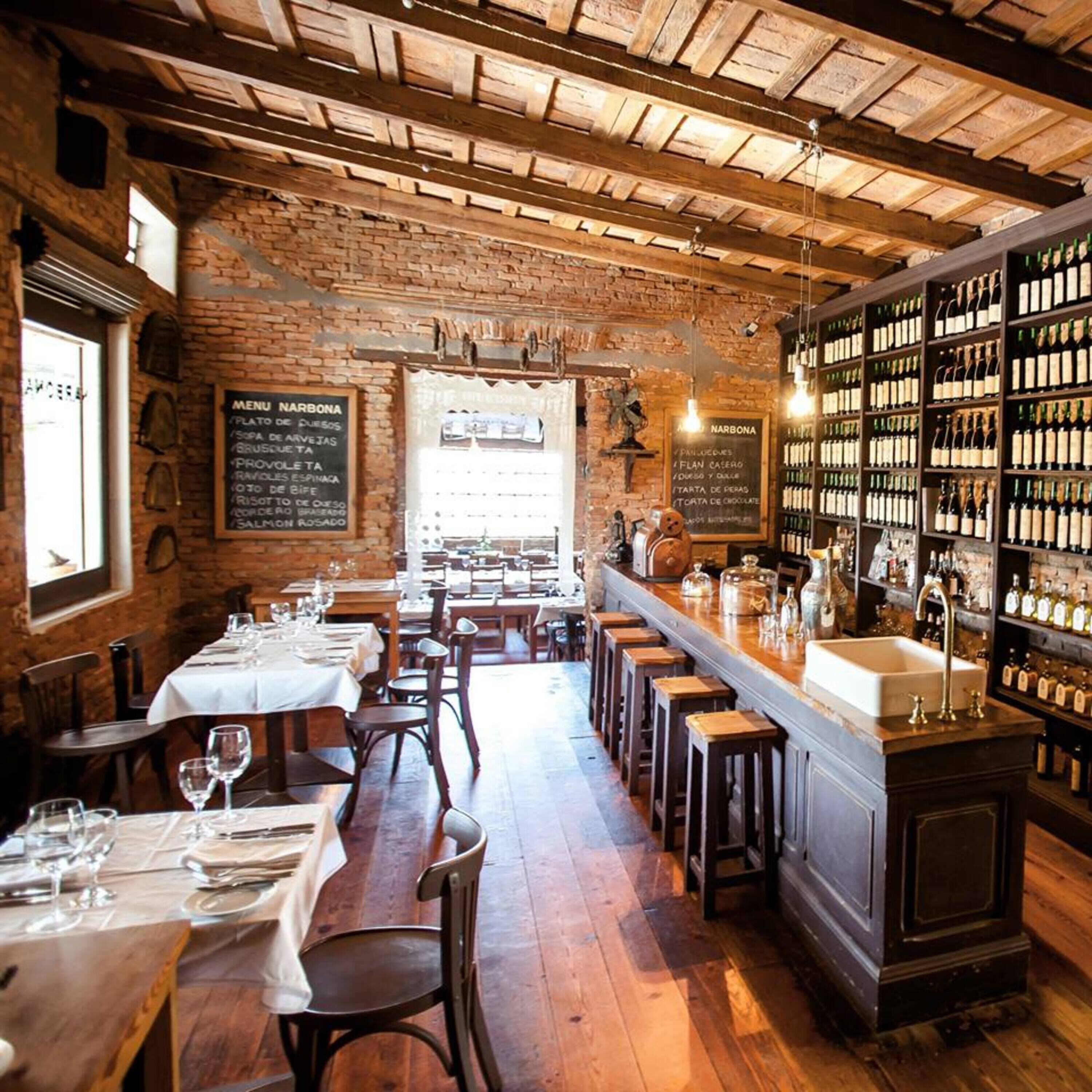 Interior rústico de un restaurante con paredes de ladrillo, vigas de madera y una larga barra con botellas de vino. Las mesas están preparadas con manteles blancos y copas de vino.