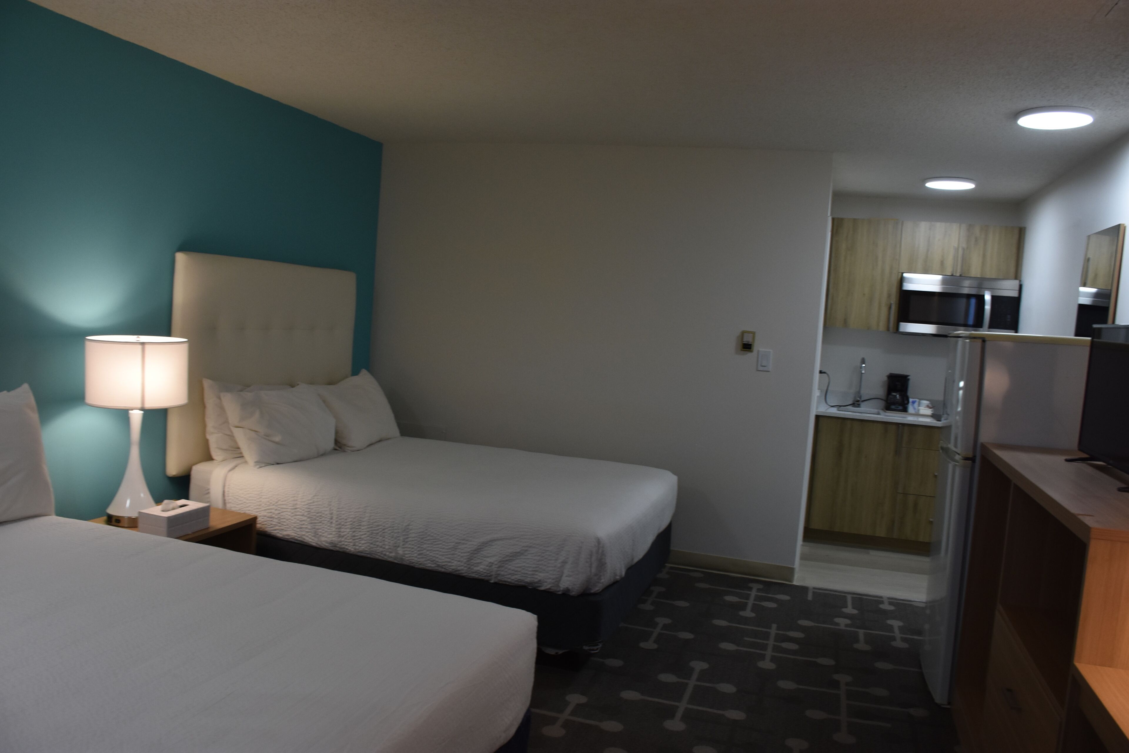 Une chambre d’hôtel avec deux lits, une table de chevet avec une lampe, une petite kitchenette équipée d’un four à micro-ondes et un téléviseur posé sur une commode en bois.