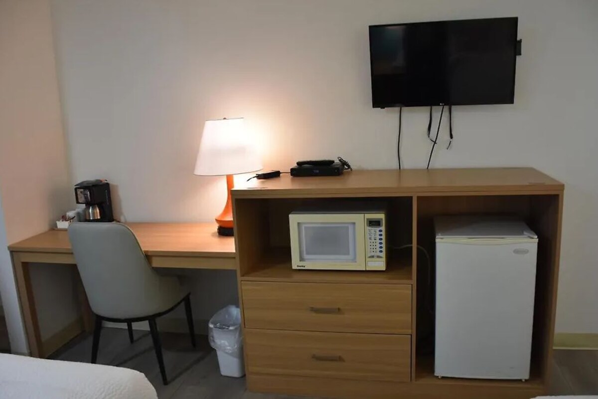 Habitación de hotel con escritorio de madera, televisor montado en la pared, microondas, refrigerador minibar, silla y mesita de noche.