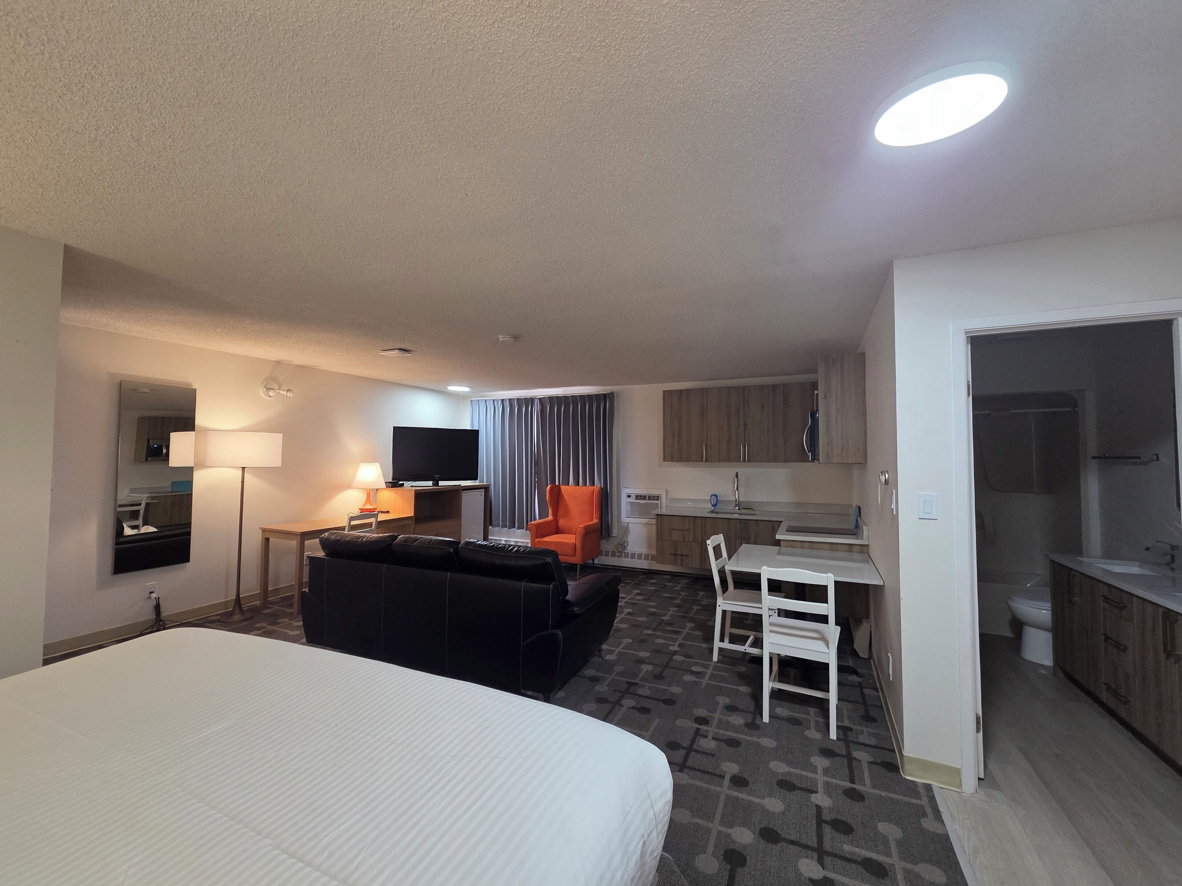 A hotel room with a bed, a sofa, a TV, a desk, a chair, a small kitchen area, and a bathroom.