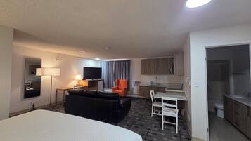 A hotel room with a bed, a sofa, a TV, a desk, a chair, a small kitchen area, and a bathroom.