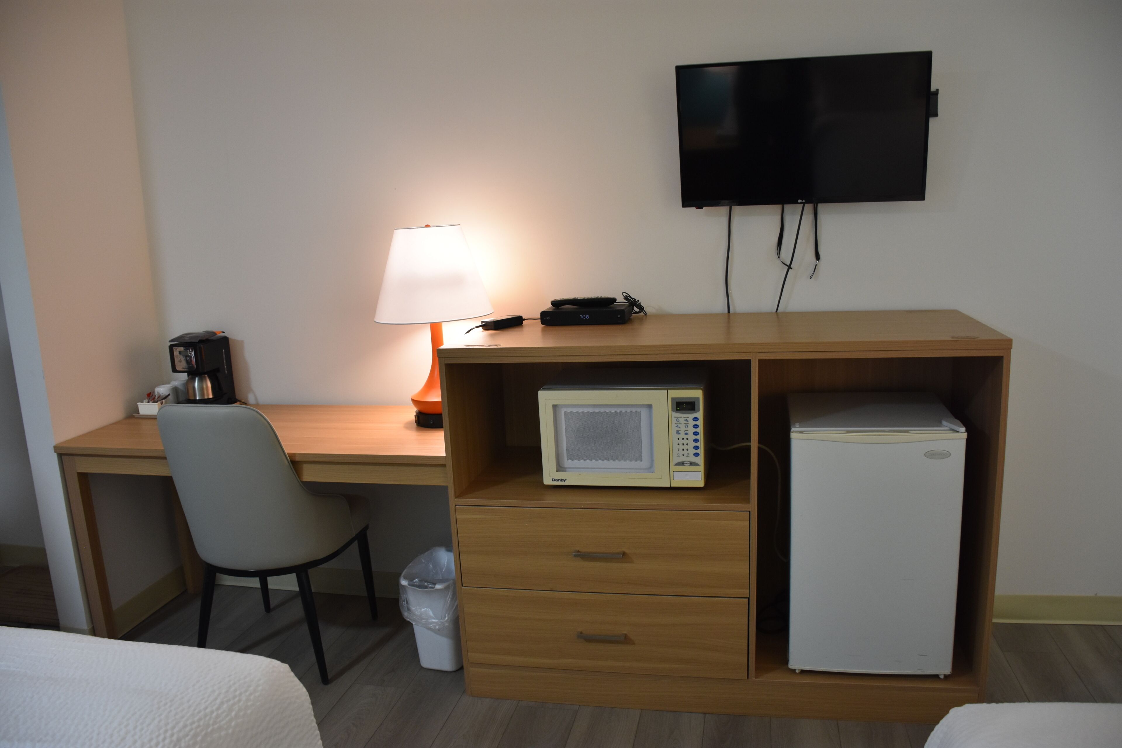 Habitación de hotel con escritorio de madera, una silla, televisor montado en la pared, microondas, refrigerador, lámpara y cubo de basura.