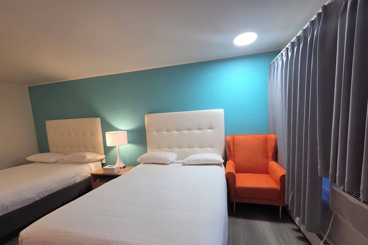 Una camera d'albergo con due letti, un comodino con una lampada, una poltrona arancione e una finestra con tende.