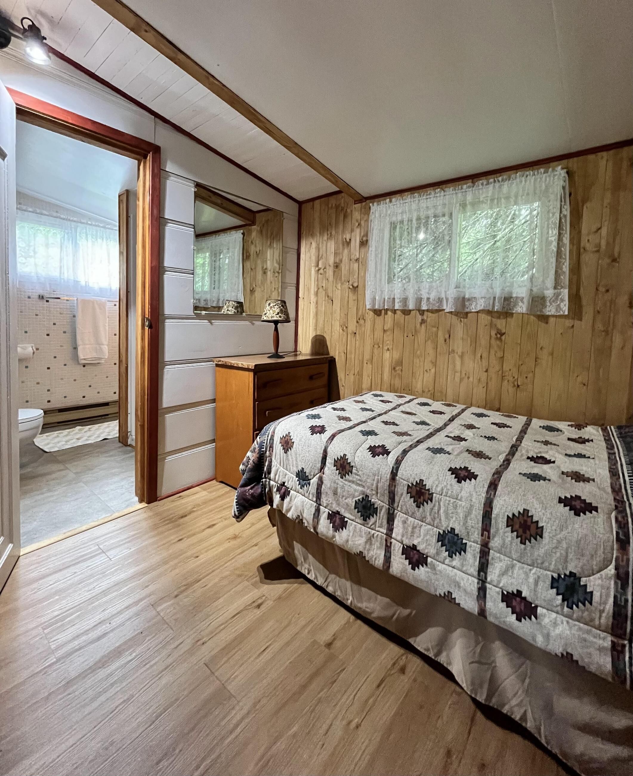 Een slaapkamer met een houten vloer, een bed met een gebloemd deken, een houten dressoir, een spiegel en een raam met dunne gordijnen.