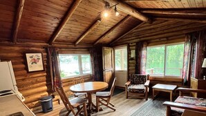 A wooden cabin interior with a dining table, chairs, a kitchen area, and large windows offering a view of the outdoors. - Kan-à-Mouche Pourvoirie Auberge et Chalets (Saint-Michel-des-Saints)
